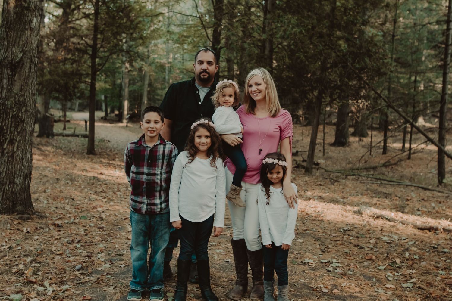 A family of five poses together on a woodland trail covered in fallen pine needles during autumn.