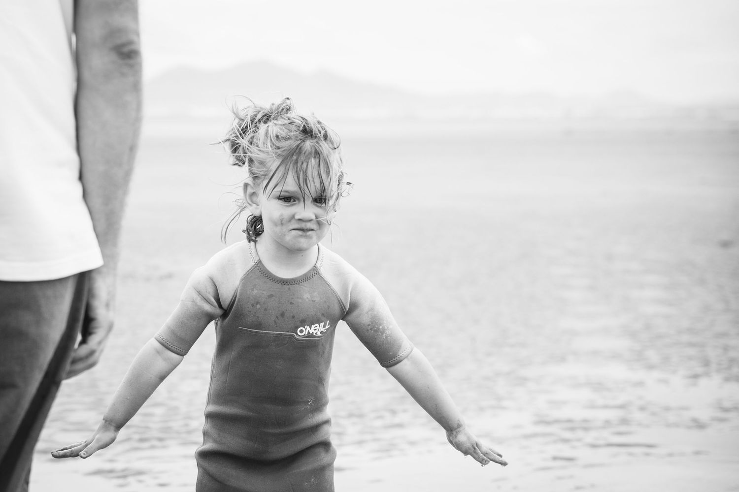 Child in wetsuit standing by ocean in dramatic black and white photo.