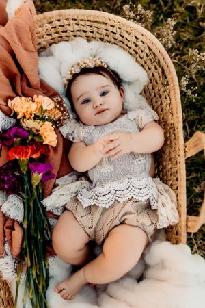 Baby in lace outfit resting in wicker chair with colorful flowers.