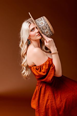 Artistic portrait featuring rust-colored dress and decorative hat against brown background.