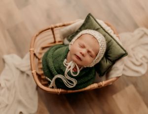 Newborn wearing white bonnet sleeping in green wrap in woven basket.