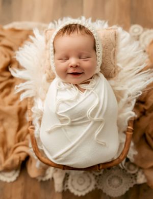 Sleeping baby wrapped in white swaddle with bonnet on textured cream blanket.