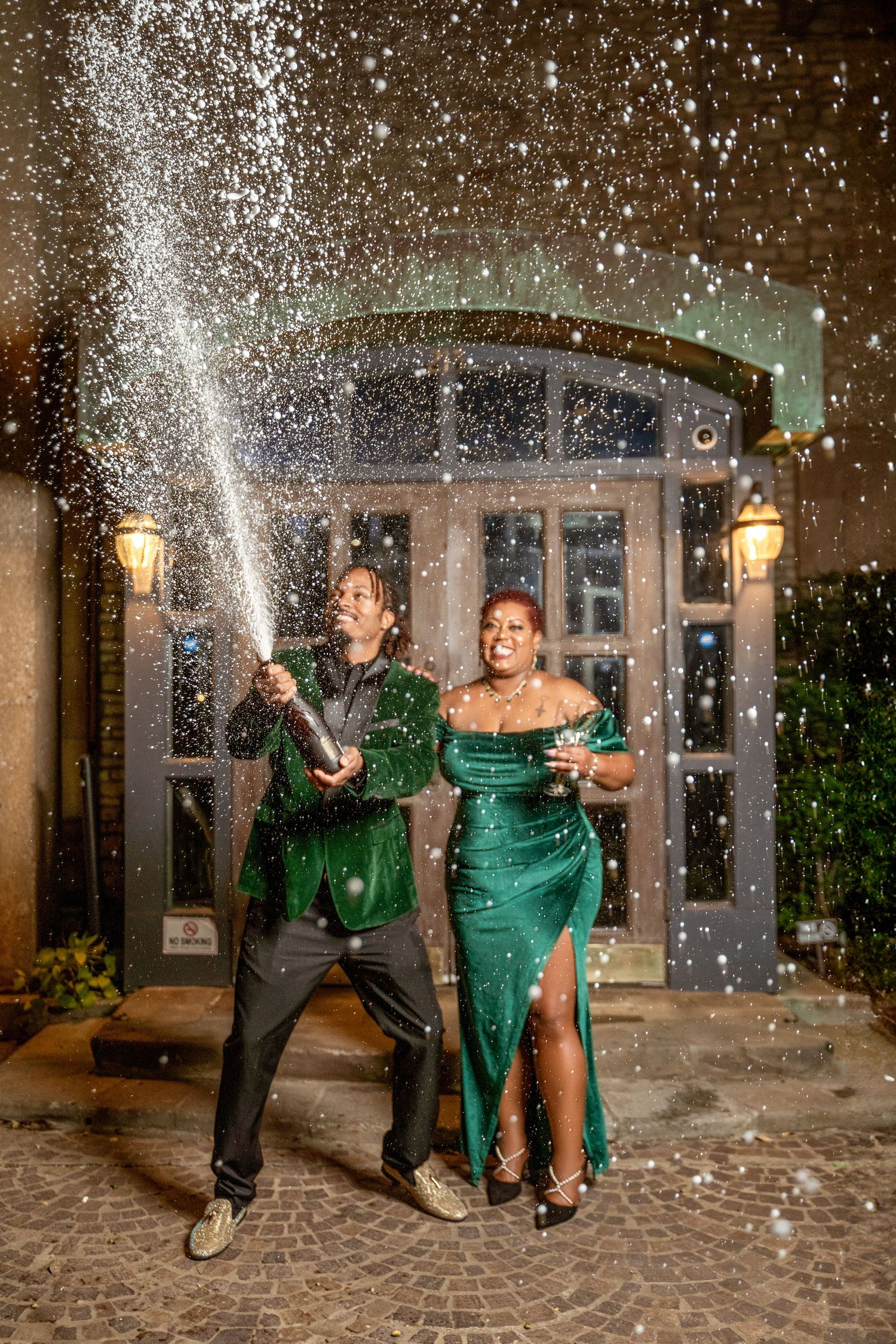 A couple in formal attire pops champagne outside during a festive celebration.
