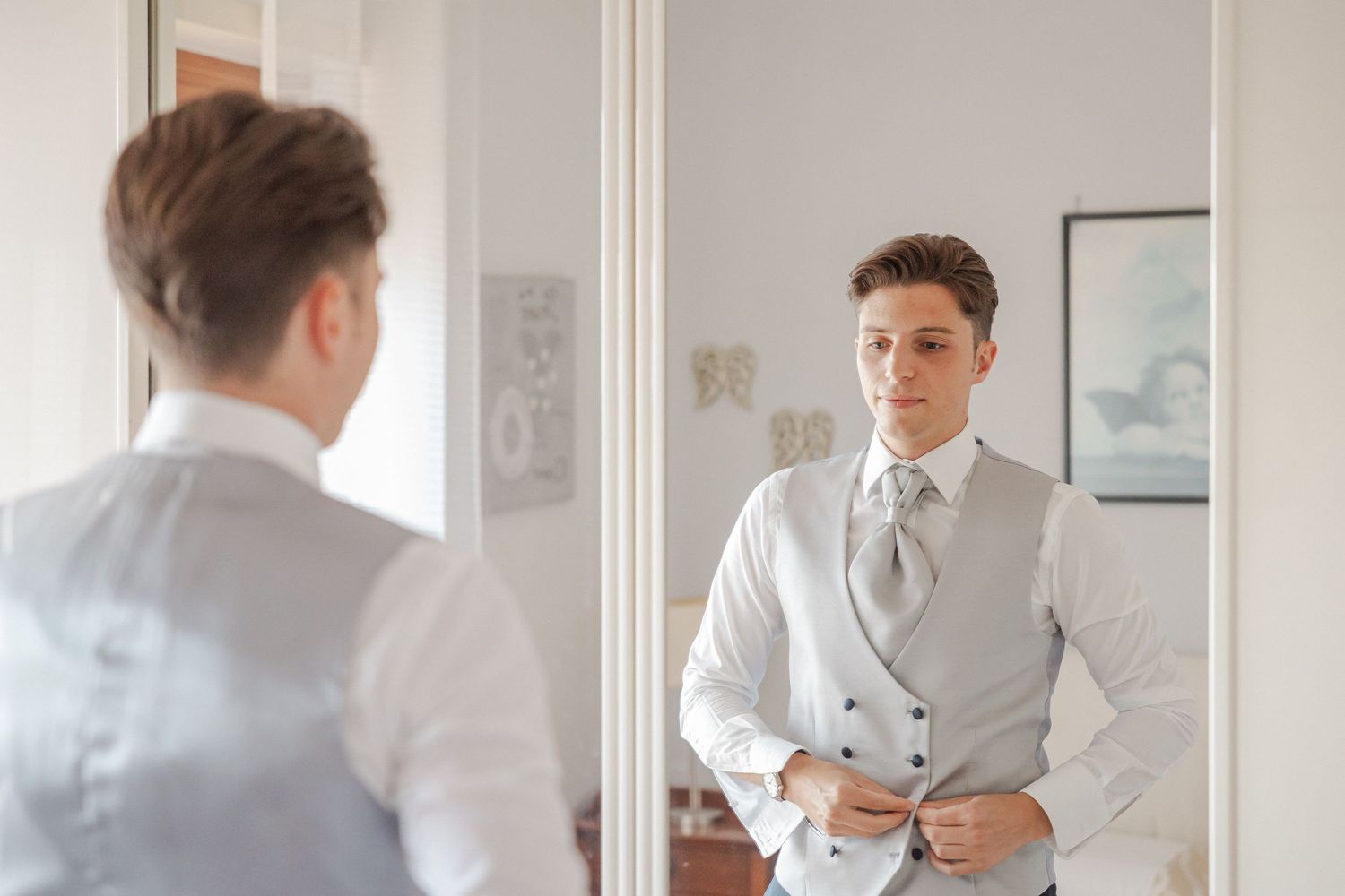 A reflection in a mirror shows someone adjusting a light gray vest and white dress shirt.