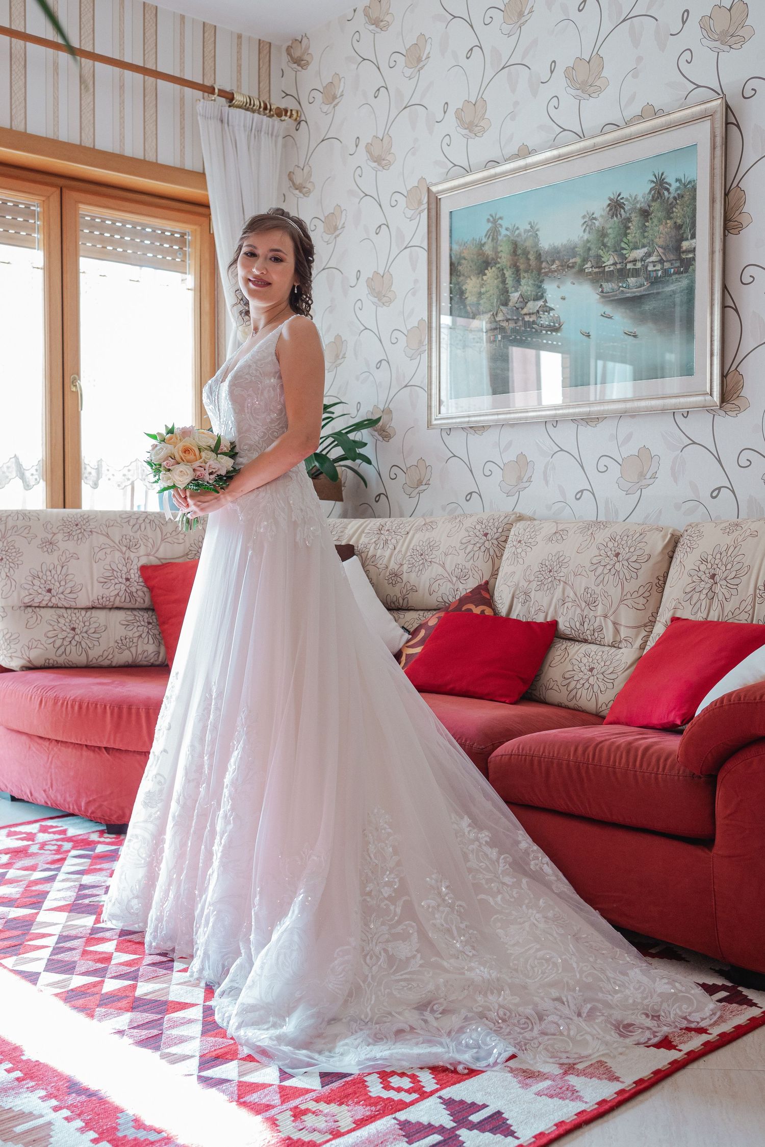 A graceful bride poses in a lace wedding gown against a floral wallpapered room with red sofa and decorative carpet.