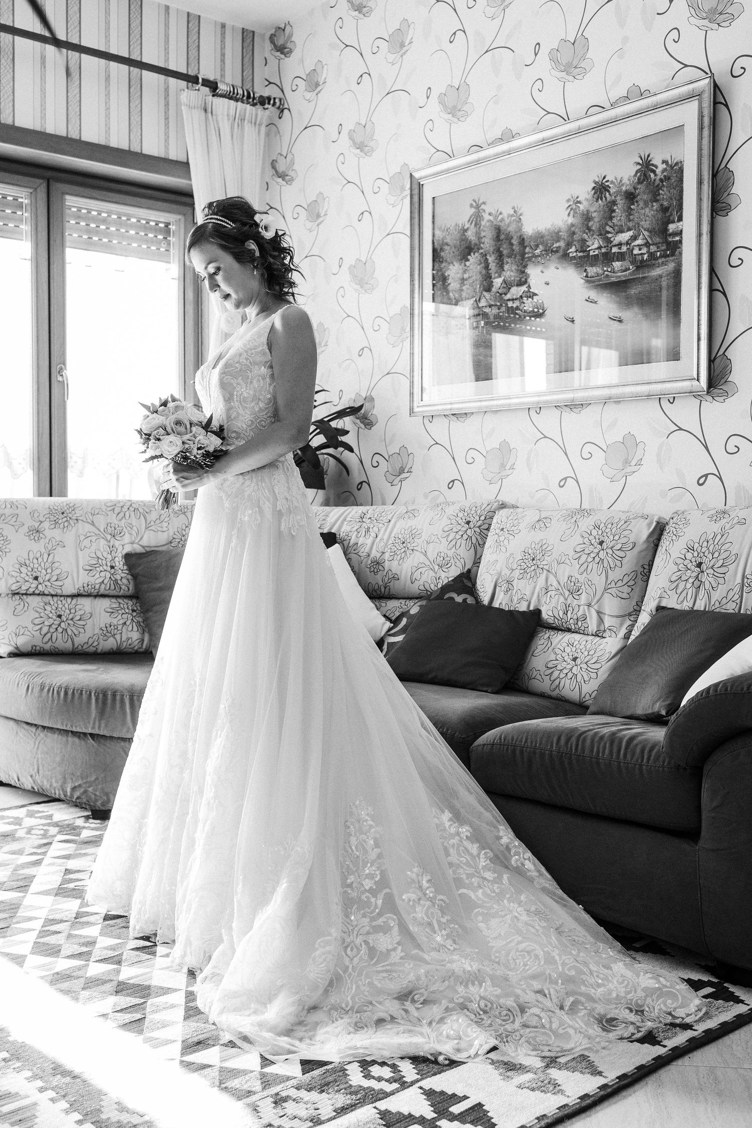Black and white photo of a bride in an elegant lace gown with train standing by a window and red sofa.