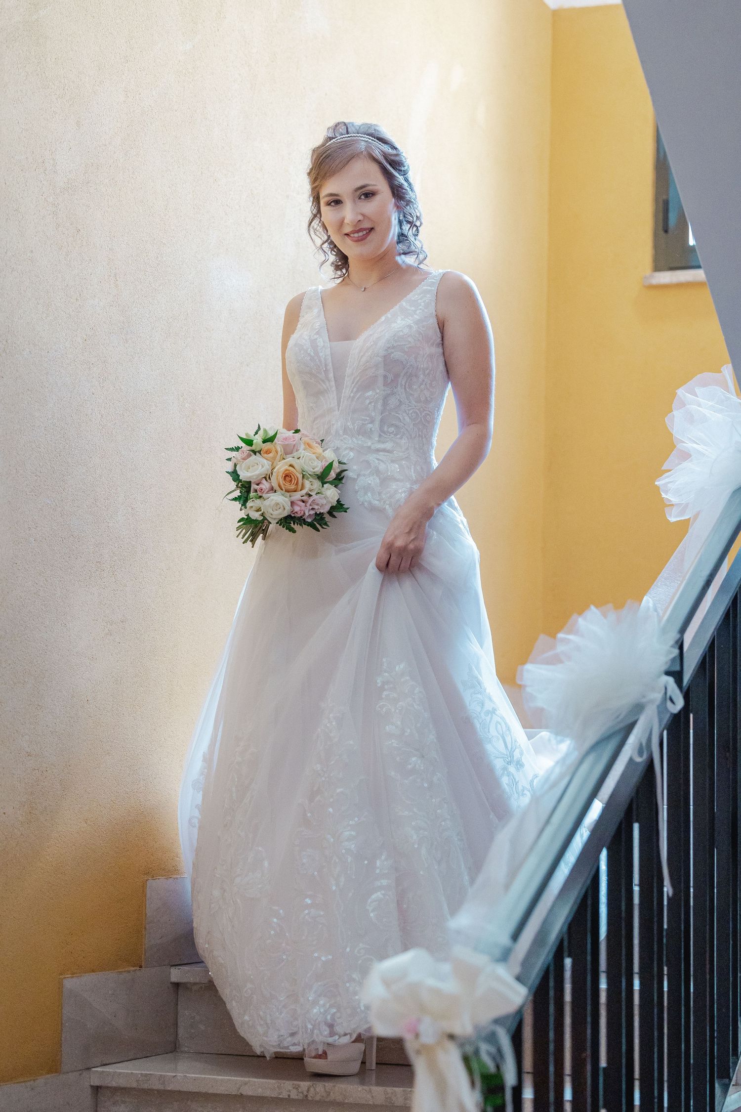 Bride in white lace wedding dress poses on staircase with peach bouquet against yellow wall.