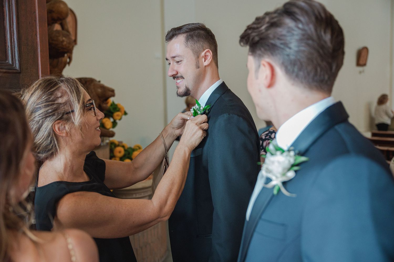 Wedding party member adjusts boutonniere on groomsman's navy suit jacket during preparation.