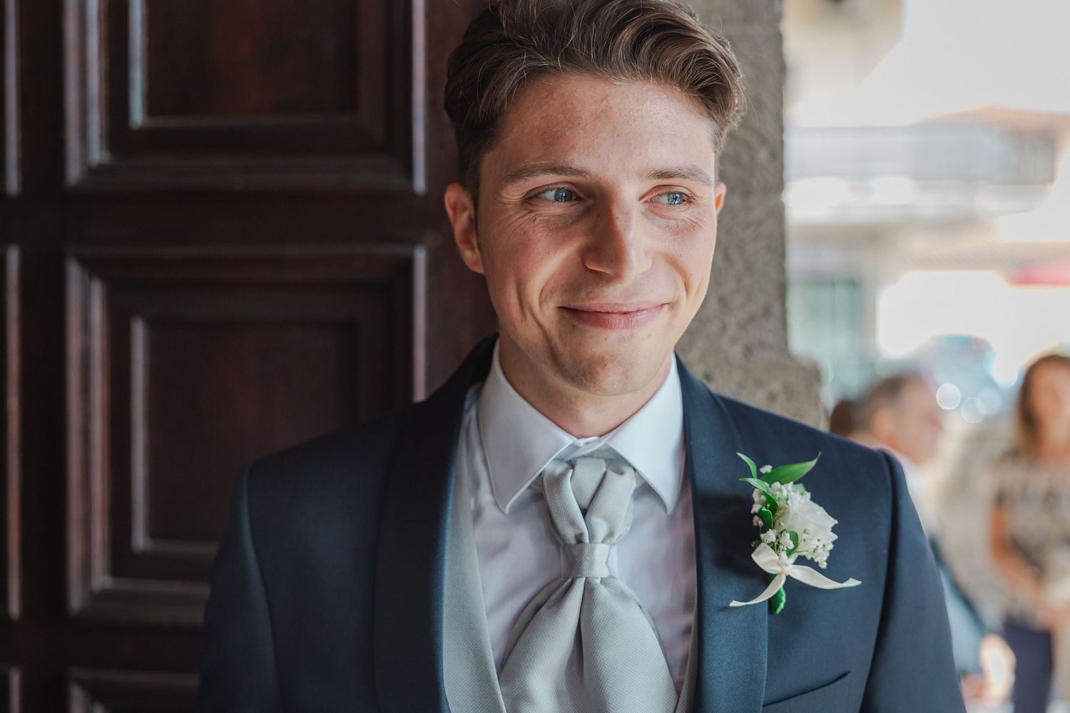 Groom in navy suit and silver tie with boutonniere smiles in natural light.