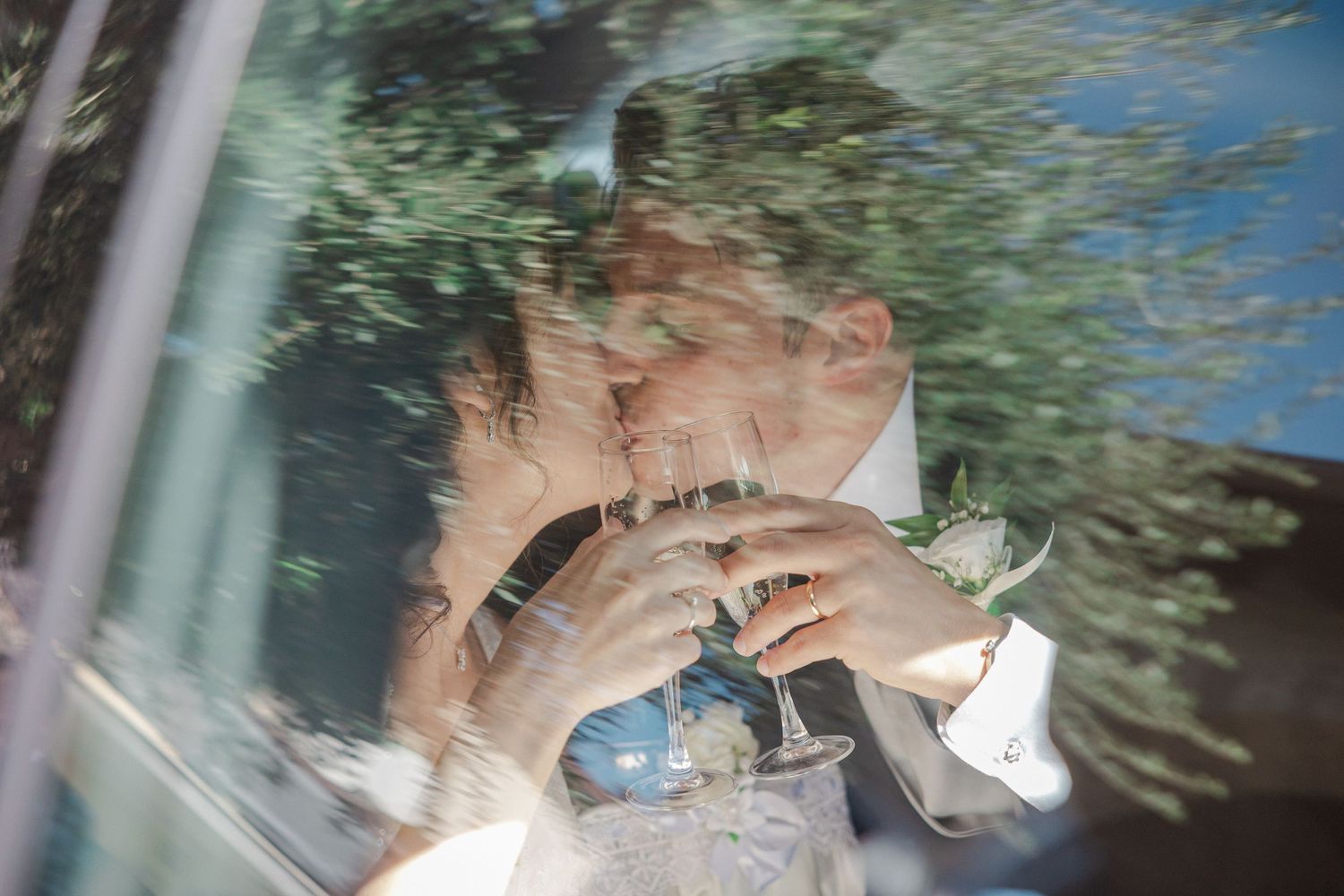 Wedding rings and hands photographed through a car window with reflections and greenery visible.