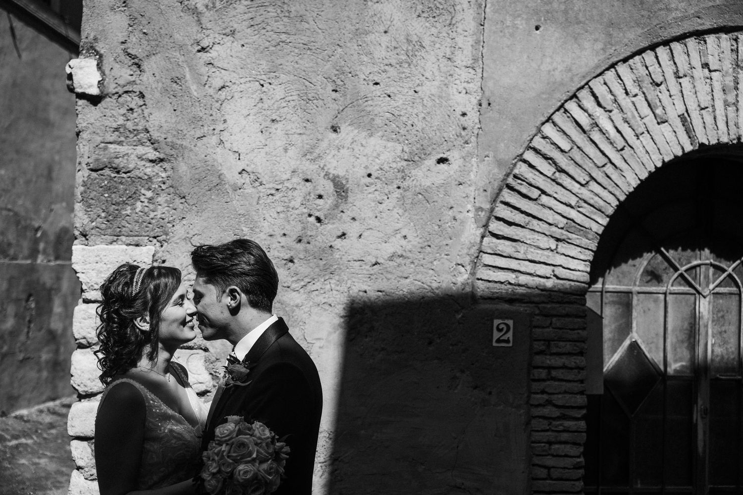 A romantic moment captured in black and white against a rustic brick archway and textured wall.