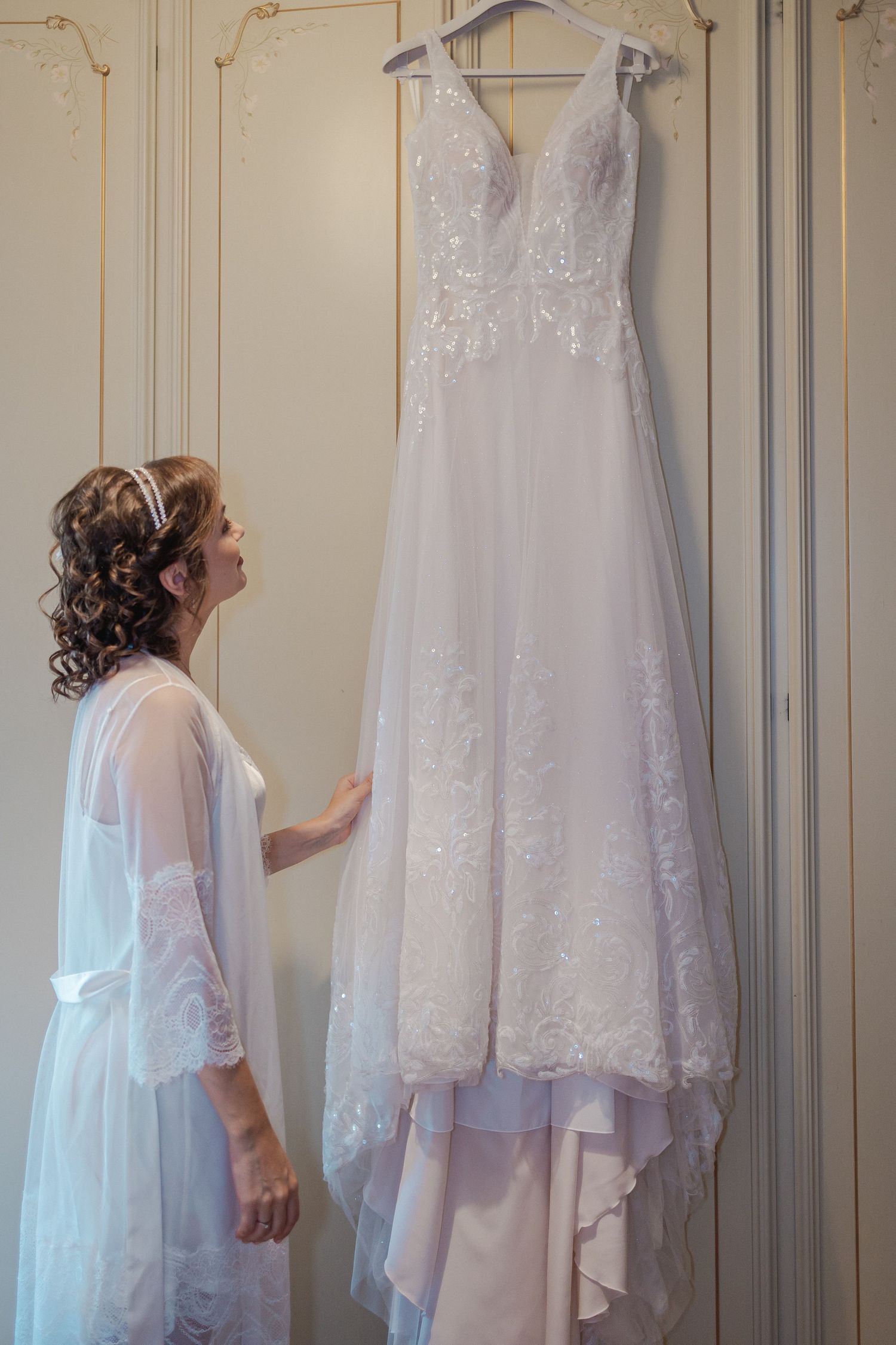 An elegant white wedding dress hangs on display against an ornate wall.