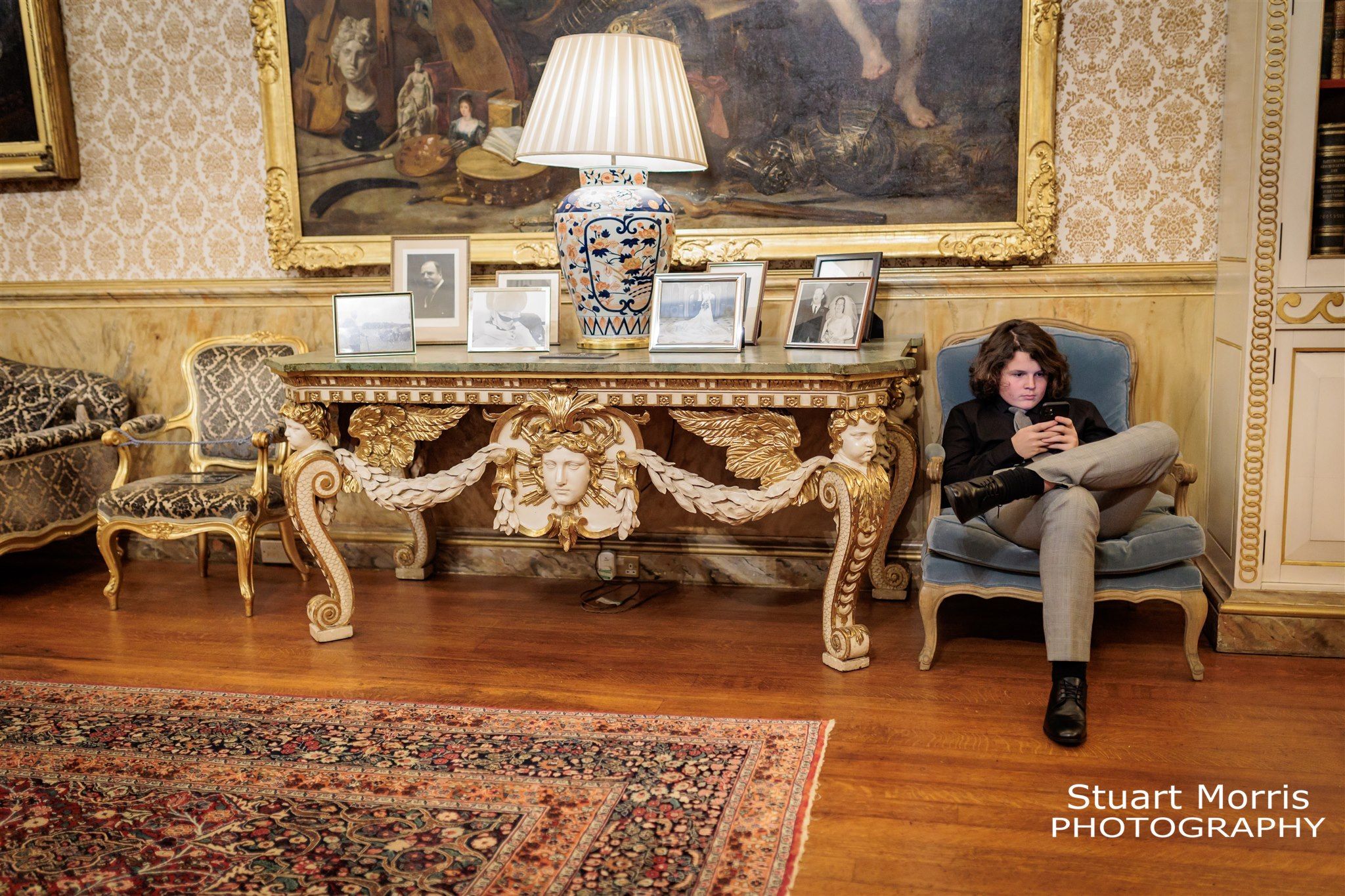 Teenage wedding guest sits on an ornate chair on his phone in the sitting room at Knowsley hall Liverpool.