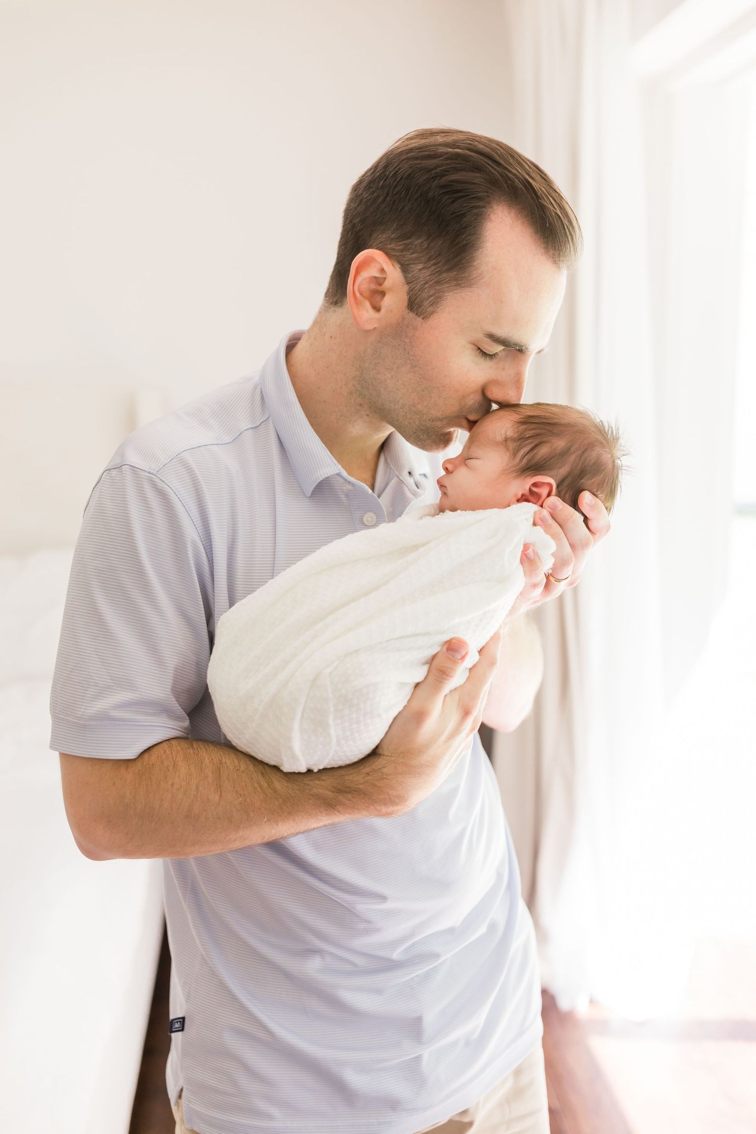 A father in grey polo shirt gently kisses his newborn baby near a bright window.