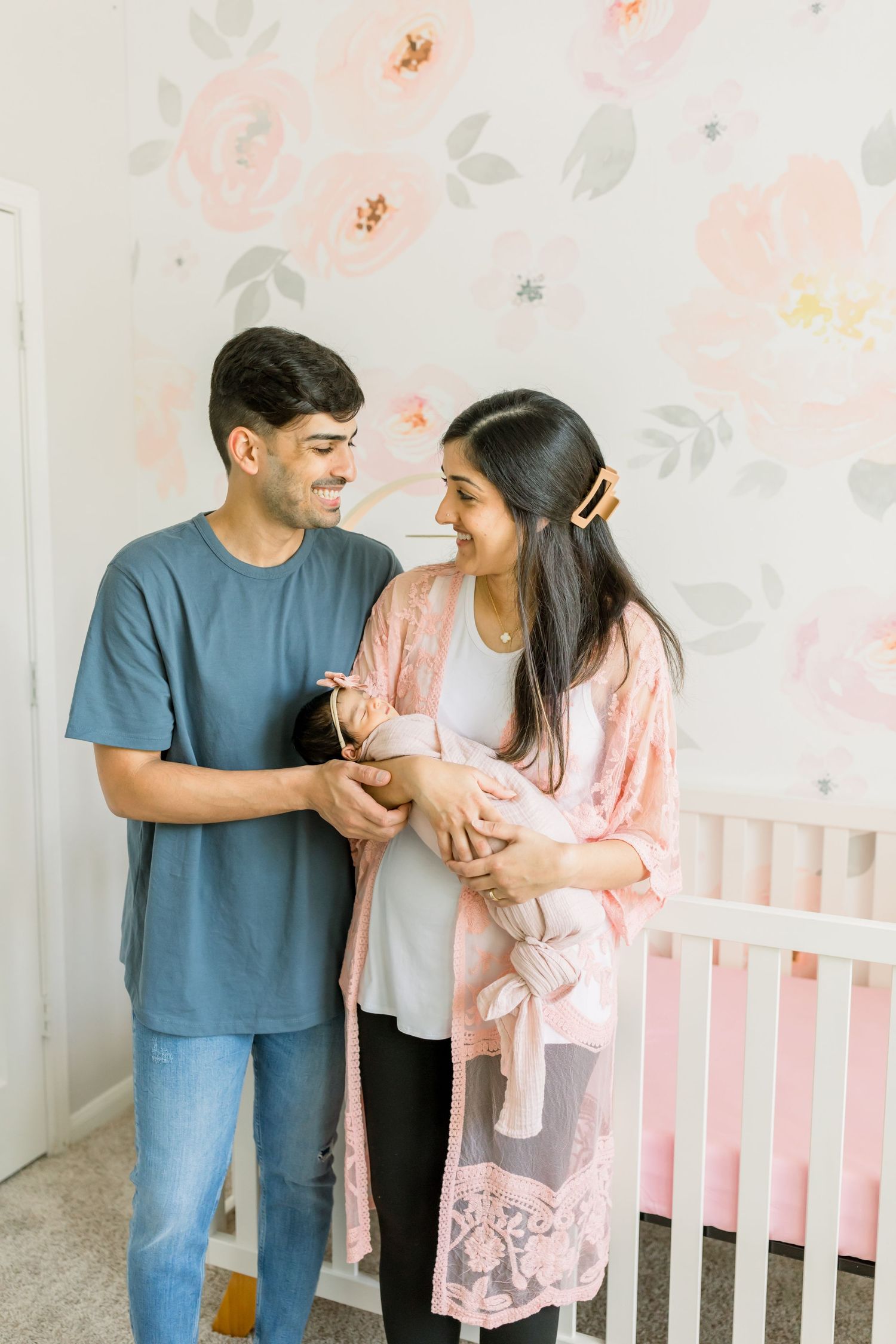 Happy parents share joyful moment while holding newborn in pink nursery setting.