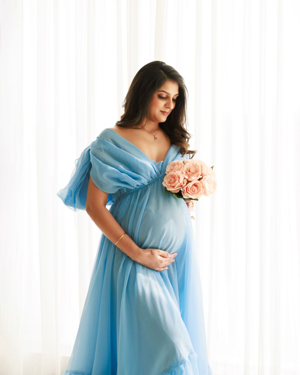 An expectant mother in a blue gown holding peachy pink roses against a white backdrop.