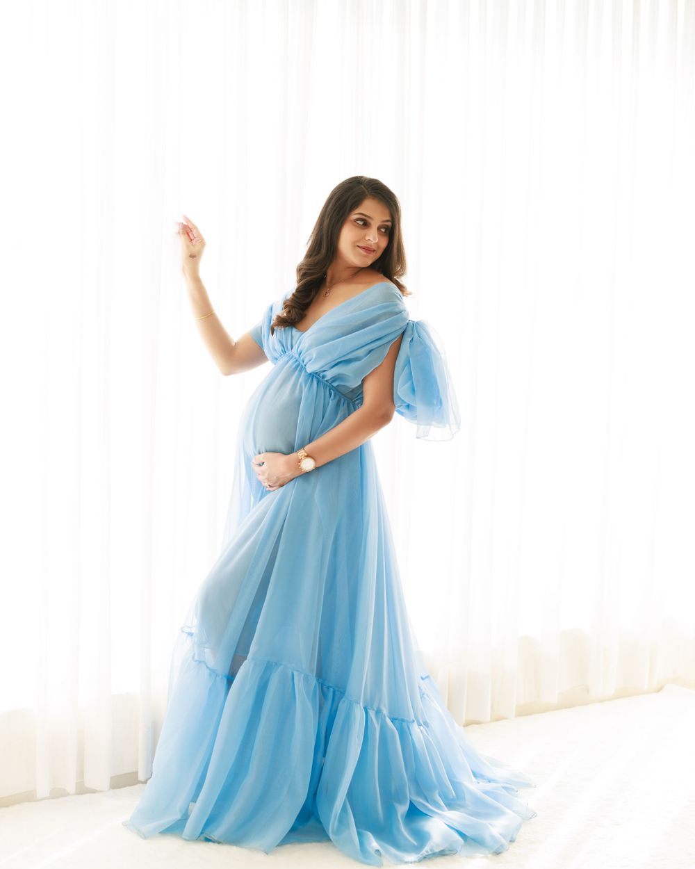 Ethereal maternity photo in flowing blue tulle gown against white backdrop.