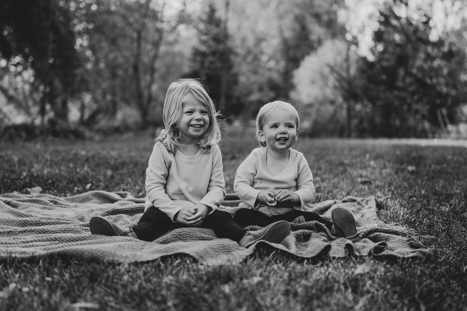 Two children share joyful laughter while sitting on a blanket outdoors in black and white.