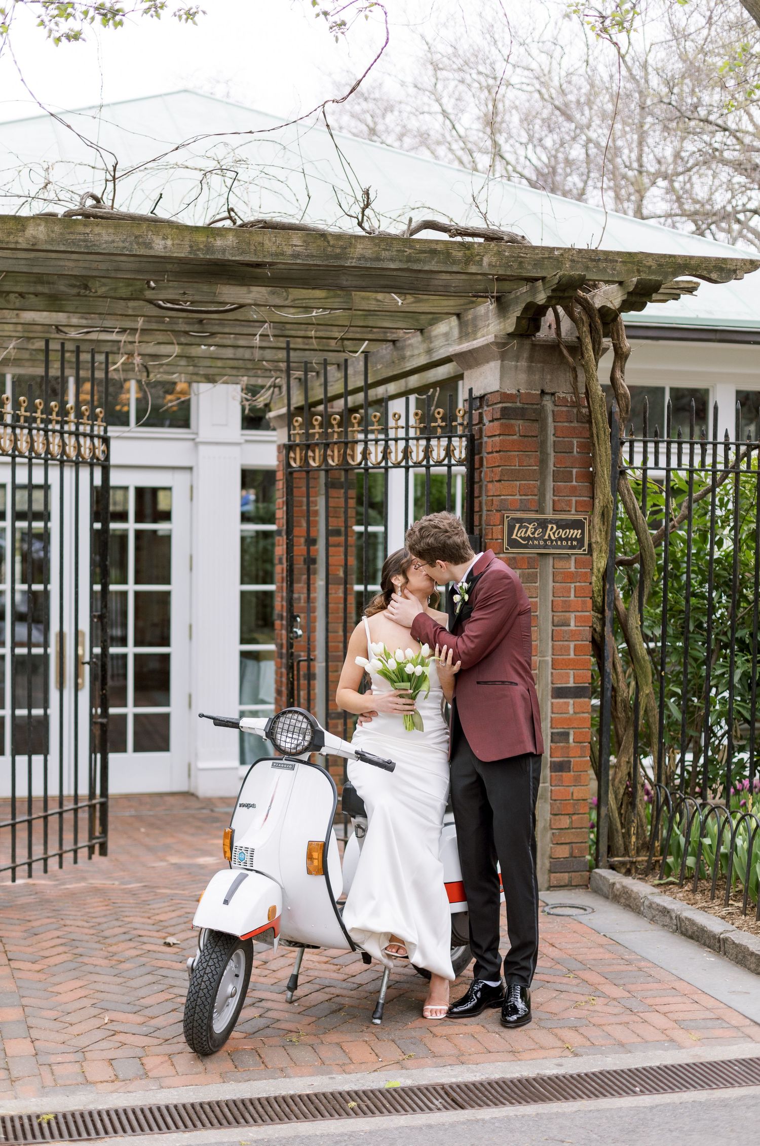 A couple in wedding attire poses with a white Vespa scooter under a brick archway.