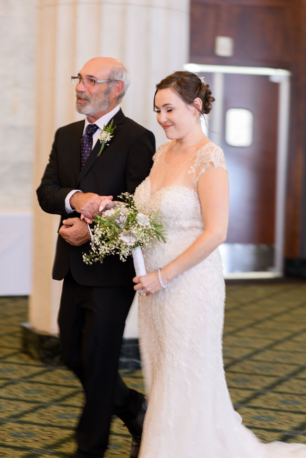 A wedding processional moment in an elegant hotel ballroom with patterned carpet.