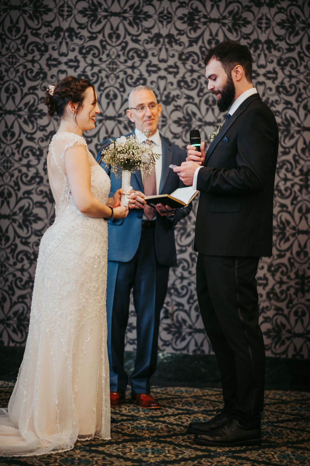Wedding ceremony in front of a damask wallpaper backdrop with floral bouquets.
