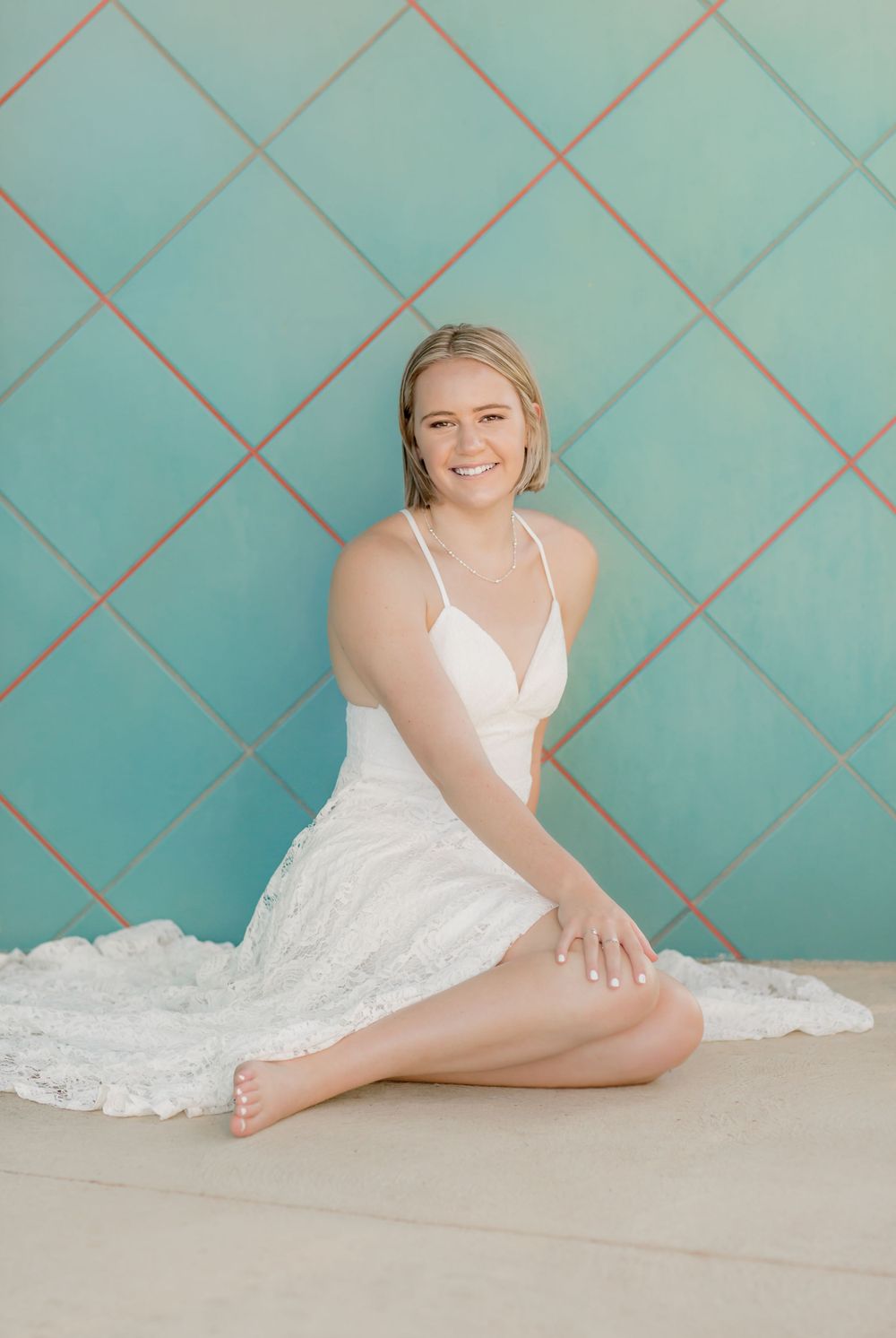 Smiling blonde woman in white lace dress sitting against teal diamond-patterned tiled wall.