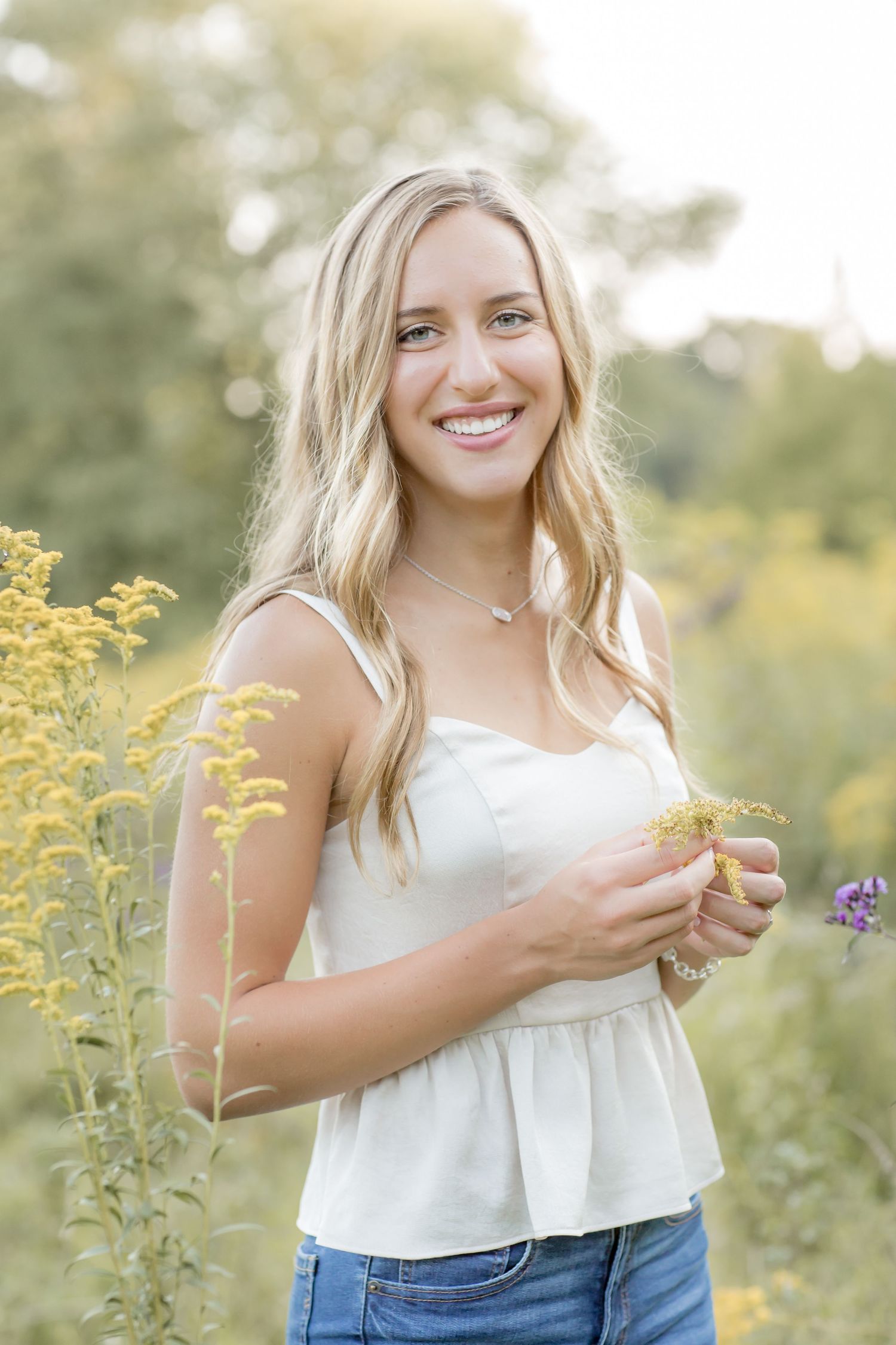 Smiling blonde teen holding wildflowers in a sunny outdoor field setting.