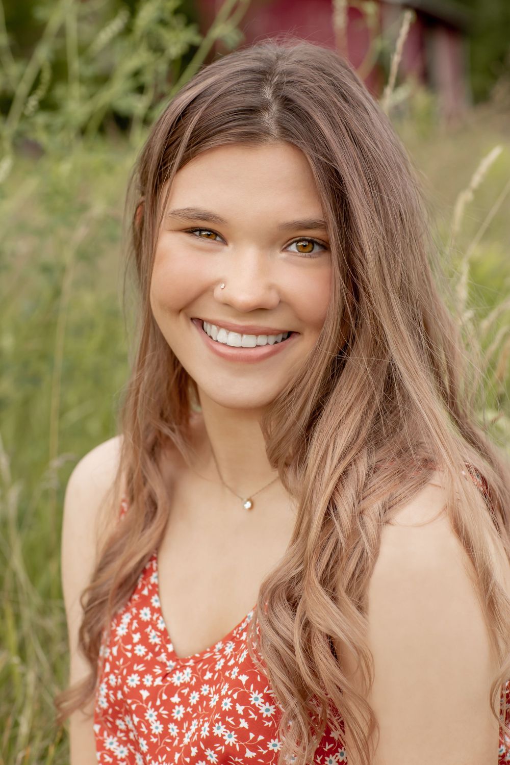 Smiling teenage girl with long brown hair in a red floral dress sitting in a field.