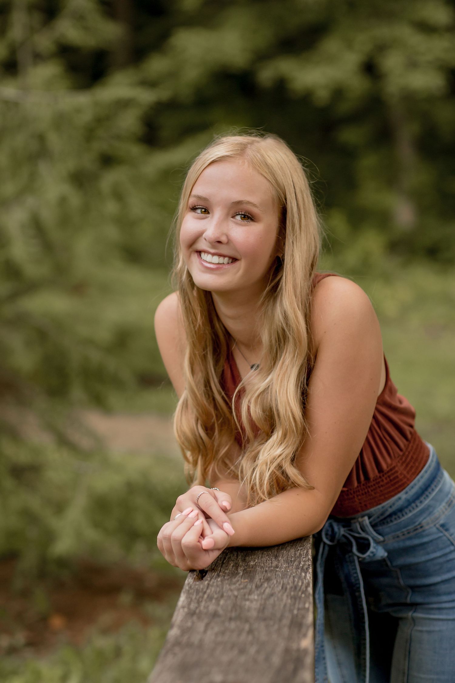 Blonde teenage girl in rust sleeveless top leaning on wooden railing in a green forest.