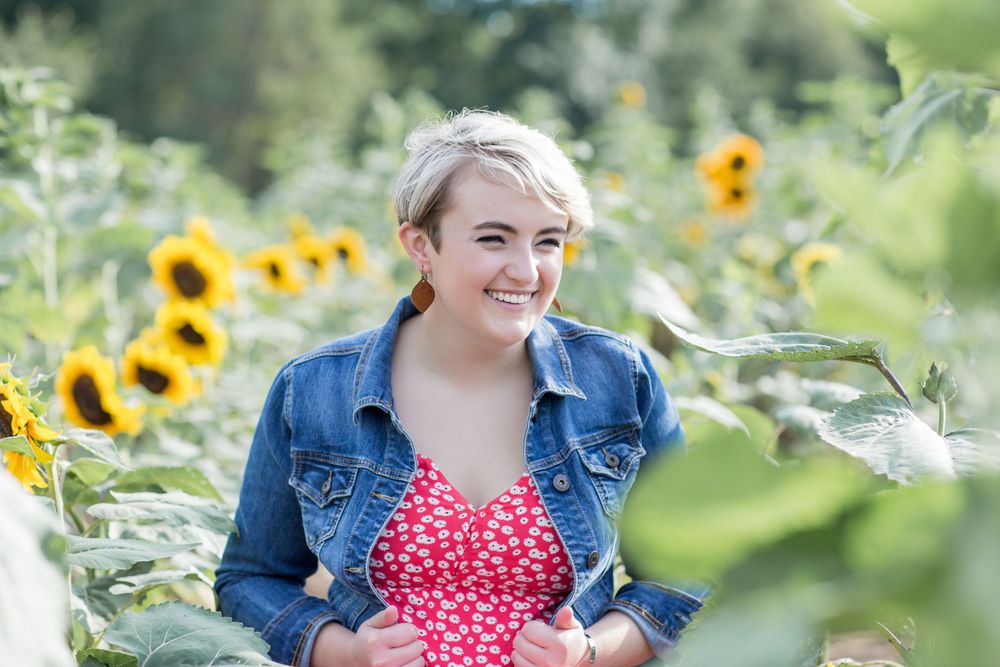 Cheerful young woman with short blonde hair in a red floral top and denim jacket in a sunflower field.