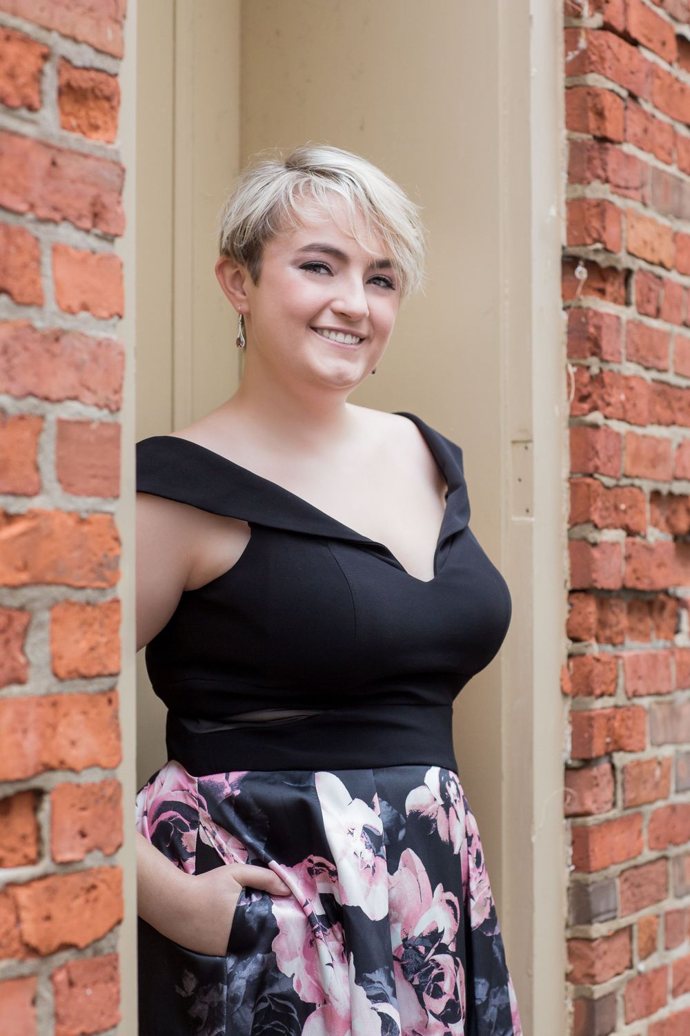 Young woman in black off-shoulder dress with floral skirt smiling in a brick doorway.