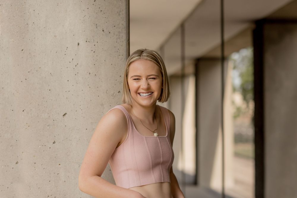 Laughing blonde woman in pink crop top leaning against concrete wall in outdoor portrait.
