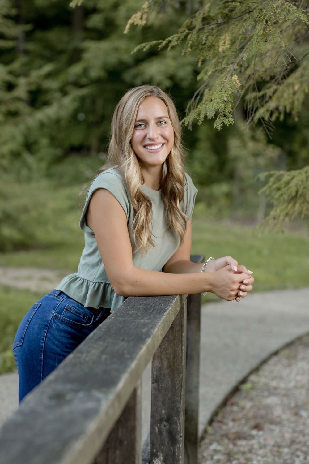Blonde teen leaning on wooden railing in green top and jeans outdoors.