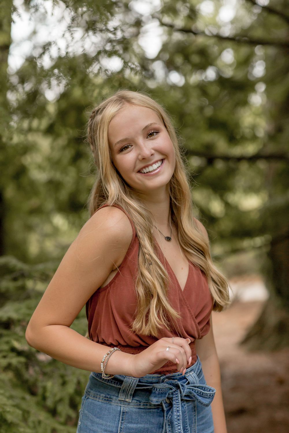 Smiling blonde teen girl in rust top and denim skirt posing on a forest path.