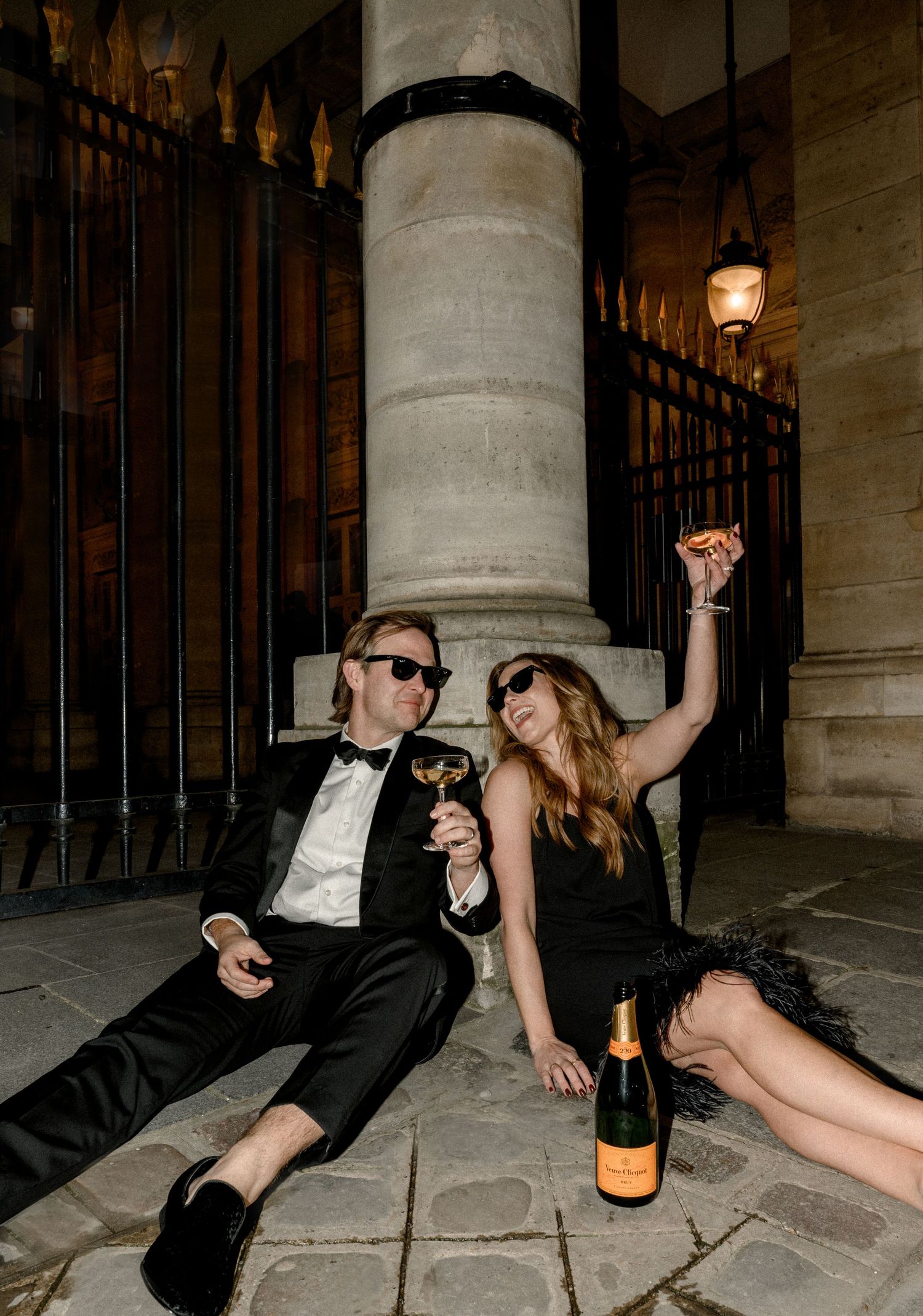 Glamorous couple lounging on stone floor with champagne in formal evening wear.