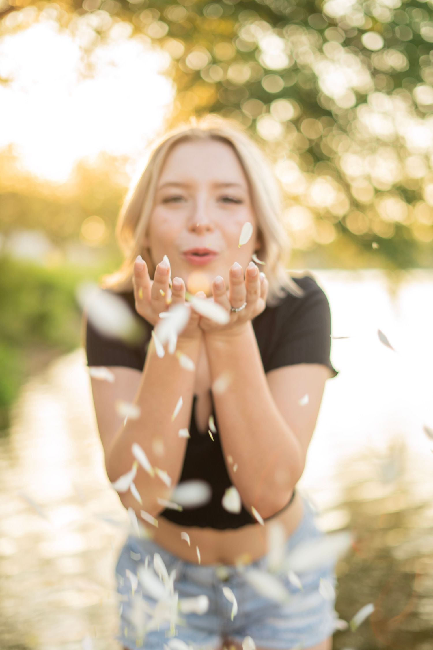 Blonde woman blowing white petals from her hands in golden sunlight near water.