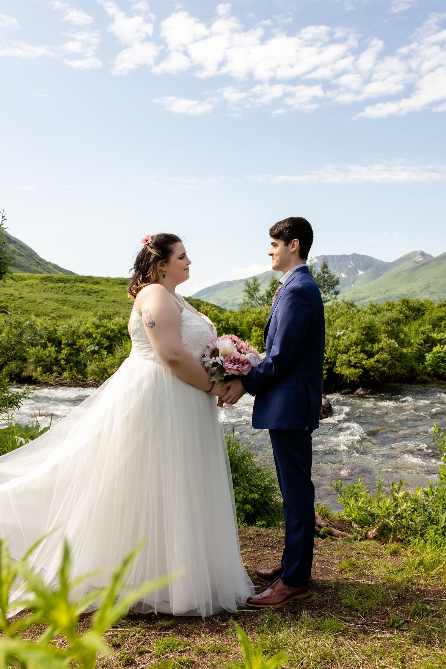 A wedding couple stands by a mountain stream with lush greenery and mountains in the background.