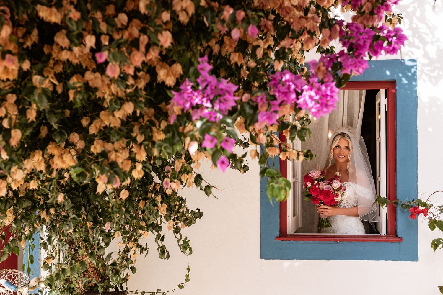 A colorful window framed by cascading pink bougainvillea and peach-colored flowers on a white Mediterranean-style wall at Quinta da Bichinha