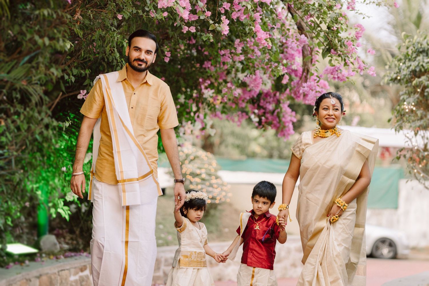 Family in traditional Indian clothing walking together beneath pink flowering bougainvillea arch.