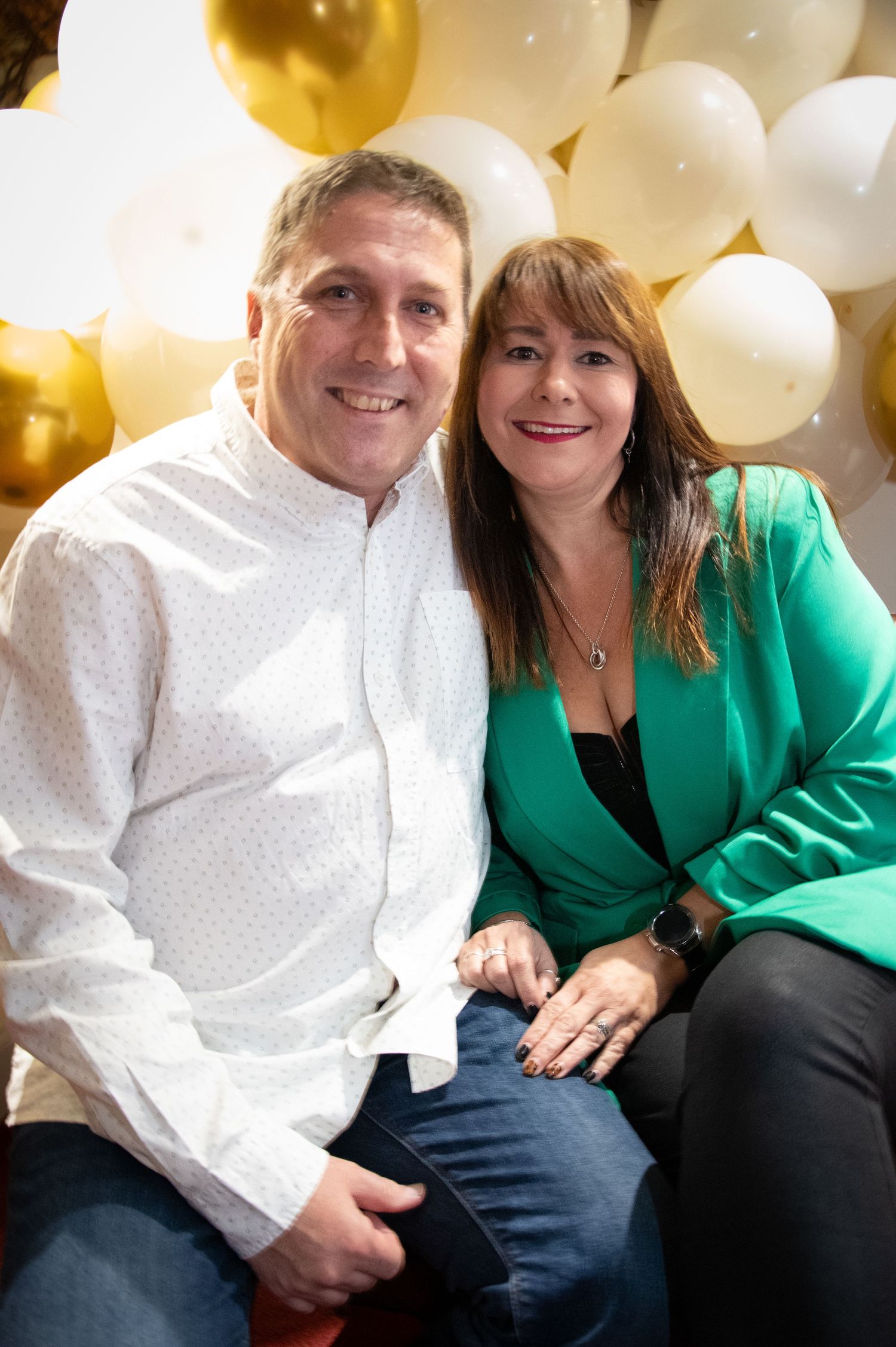 Couple in formal attire sits together against gold and white balloon display.