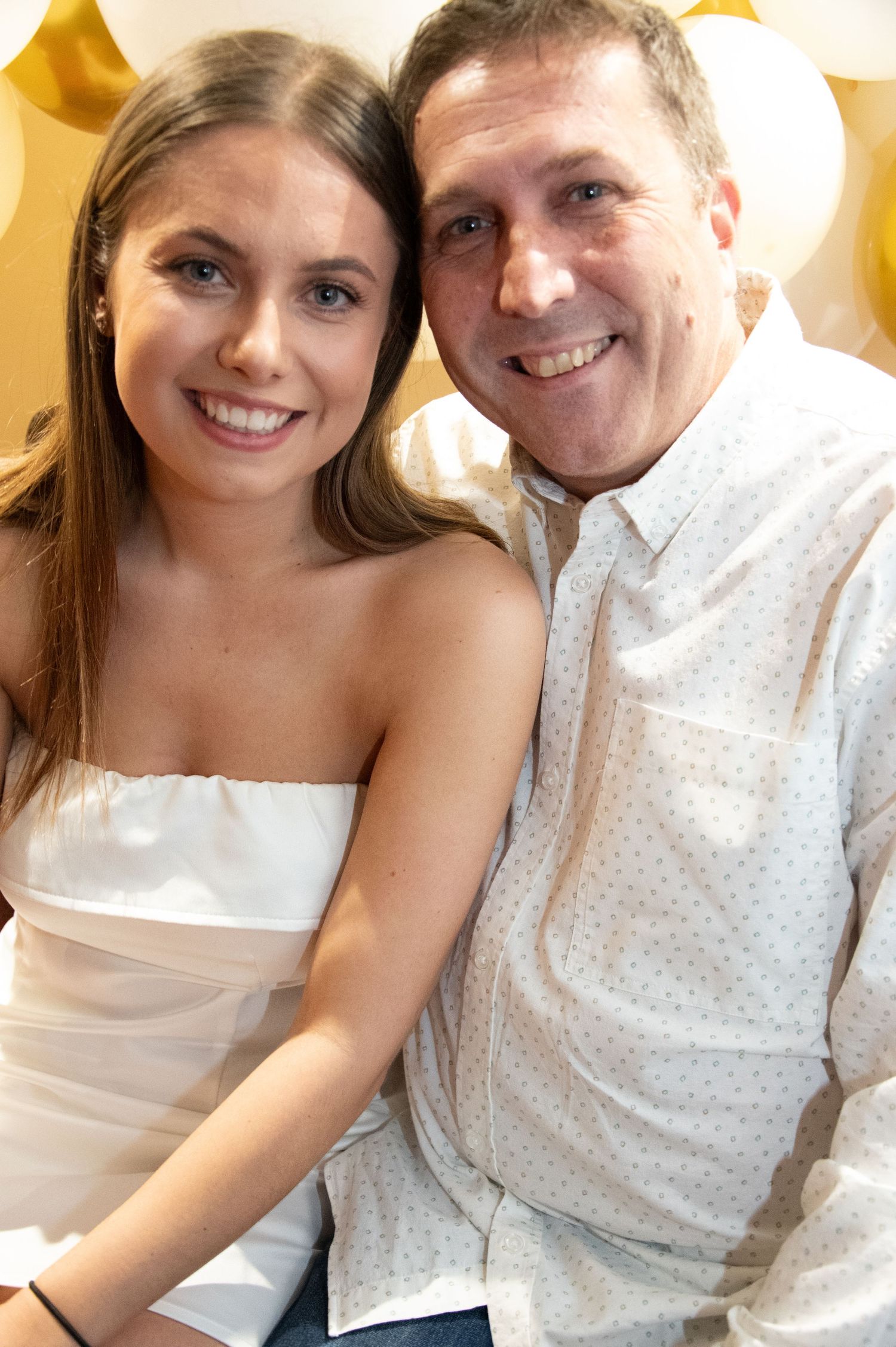 Two people pose closely together against festive gold and white balloon wall.