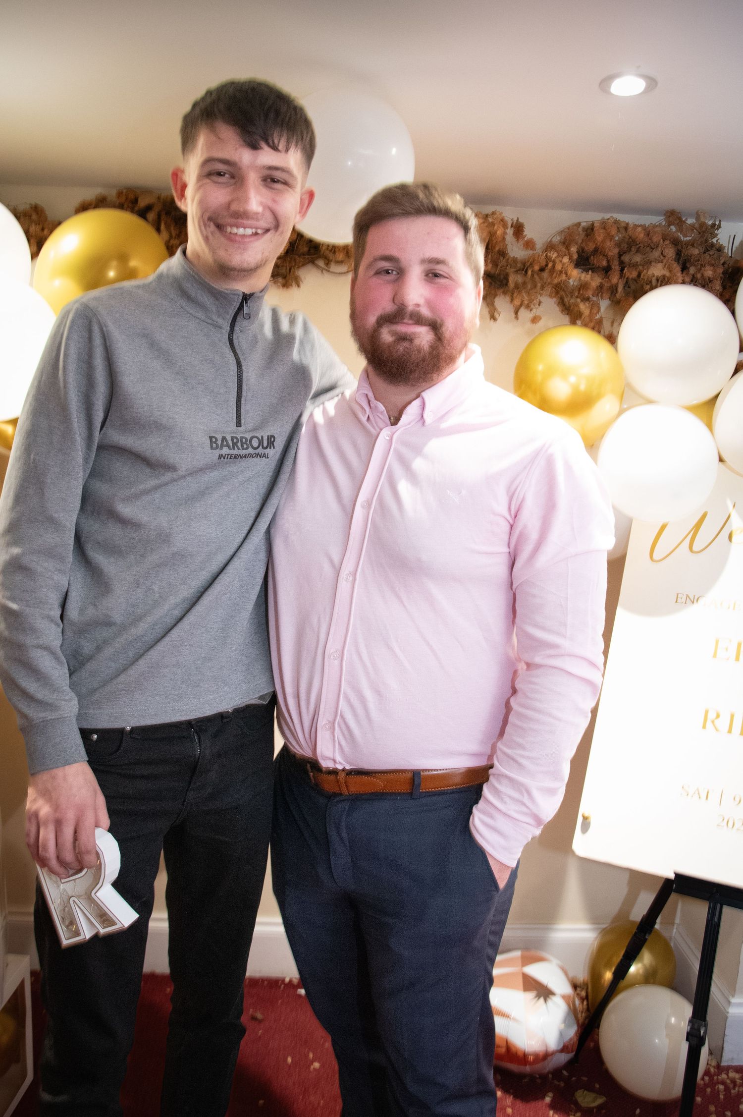 Two people smile together at a celebration with gold and white balloon decorations.