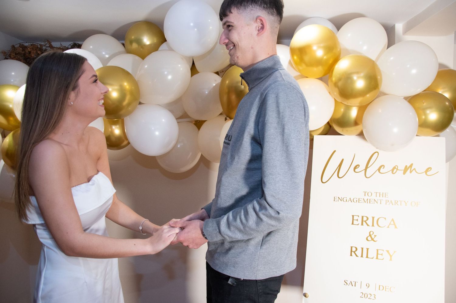 A couple shares a happy moment in front of their engagement party welcome sign and balloon wall.