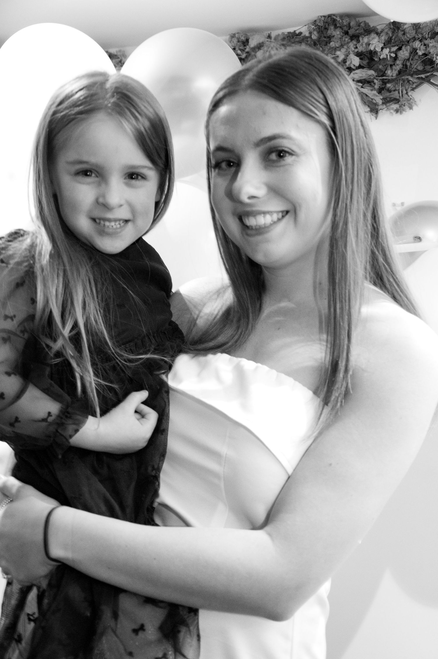 A woman in a white dress poses with a young girl in black at an event with decorative balloons.