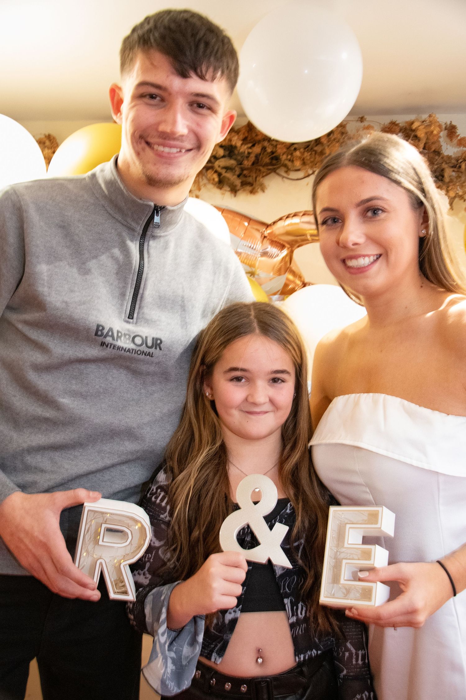 Three people pose together holding silver letters with gold and white balloons in the background.