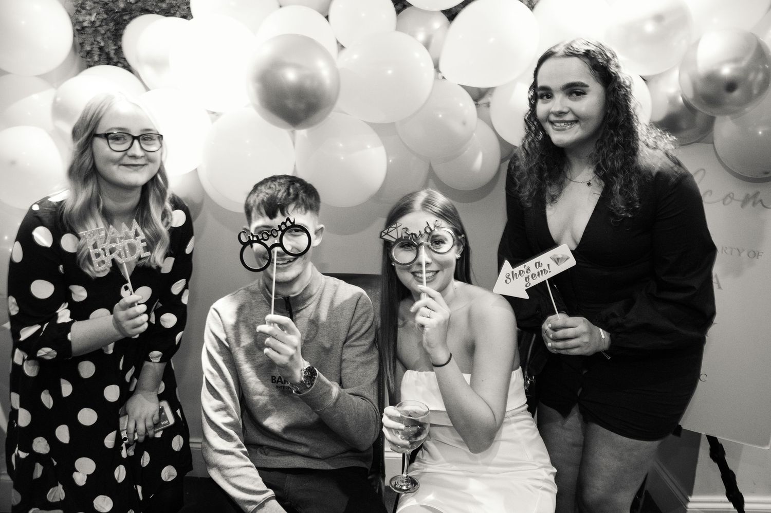 A group poses with photo props against a backdrop of white and gold balloons.