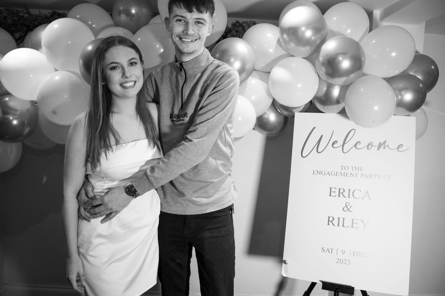 A couple poses together at their engagement celebration with white and gold balloon decorations.