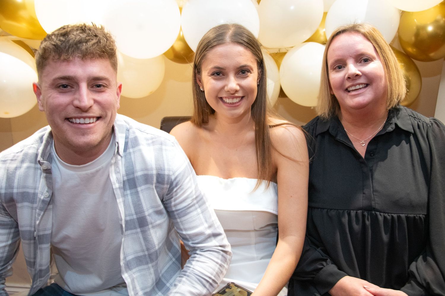 Three people smile together at celebration with gold and white balloon decor.