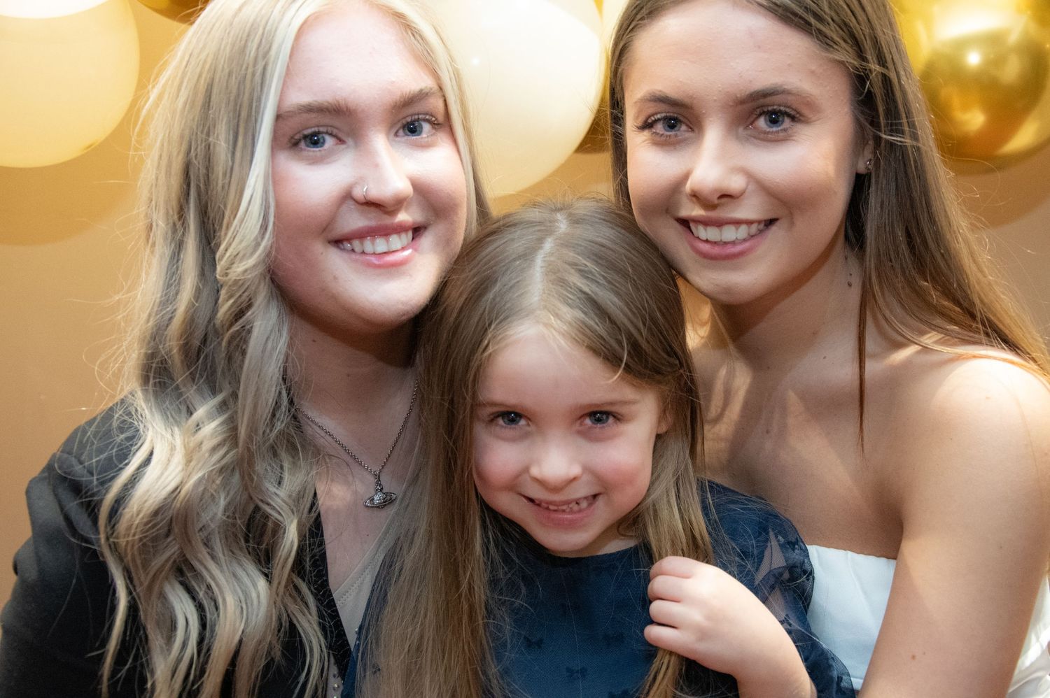 Three smiling figures pose closely together with gold balloons in background.