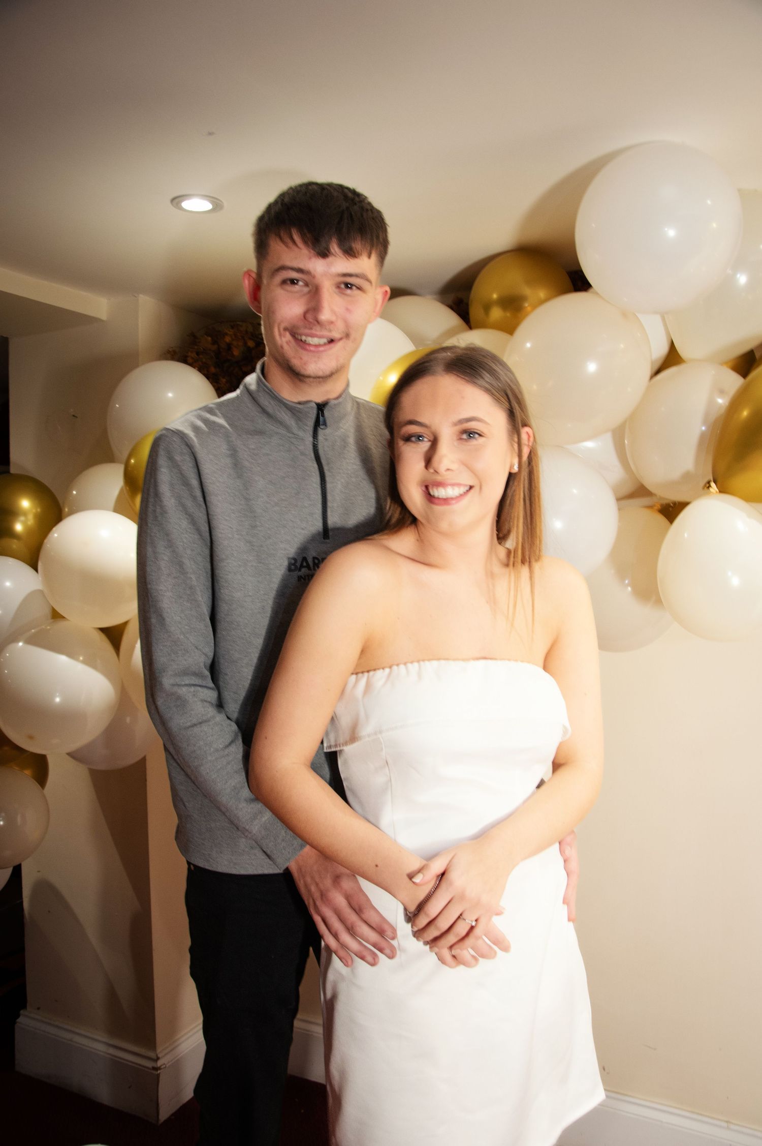 Couple stands together at celebration with white and gold balloon backdrop.